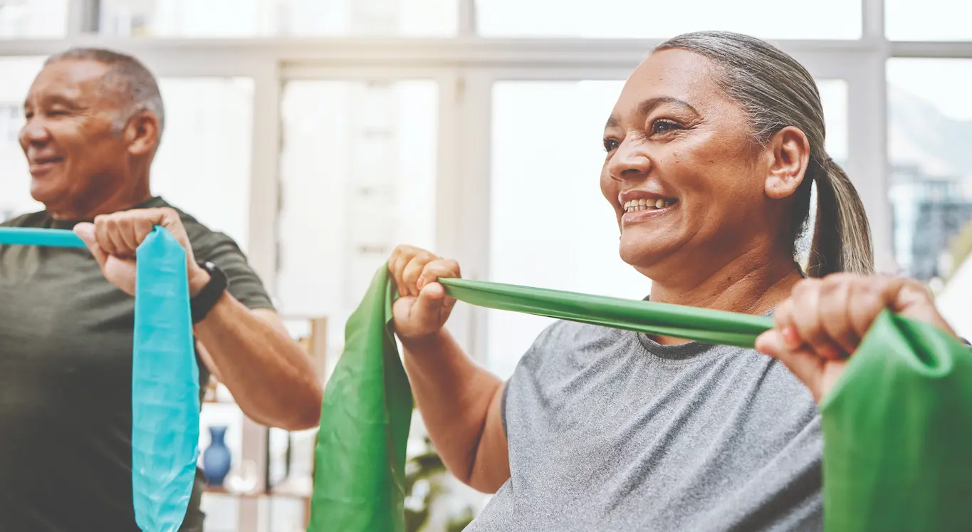Two older adults smiling while exercising with resistance bands indoors, bathed in natural light from large windows behind them.