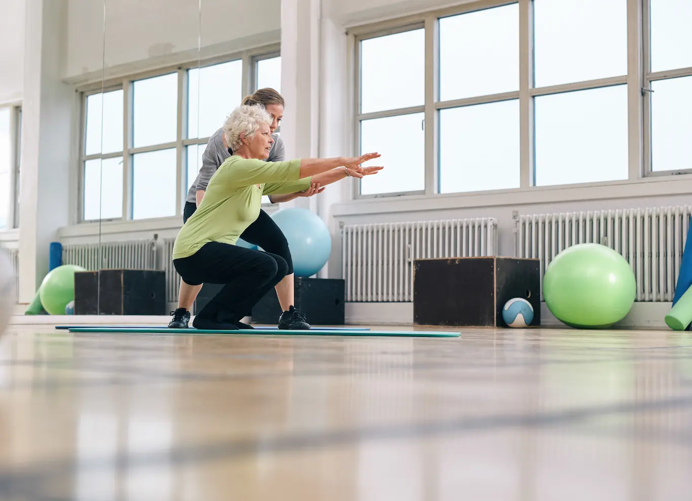 An older woman performs a squat on a yoga mat in a gym, with a younger instructor supporting her. Exercise balls and gym equipment are visible in the background.