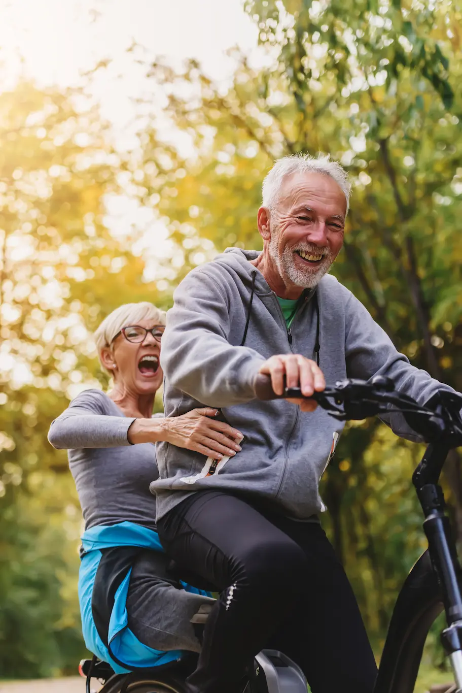 An older man and woman, both smiling and laughing, ride a bicycle together outdoors on a sunny day, surrounded by green trees and warm light.