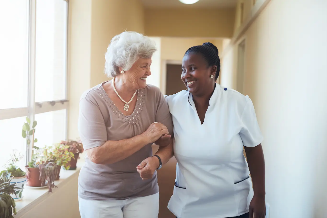 An elderly woman and her SeniorConcierge caregiver walk arm-in-arm down a bright hallway, both smiling warmly at each other. There are potted plants on the windowsill to their left, creating a welcoming scene for St.Louis Seniors.