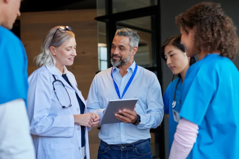 A diverse group of five medical professionals and a man in a business shirt are gathered, looking at a digital tablet.