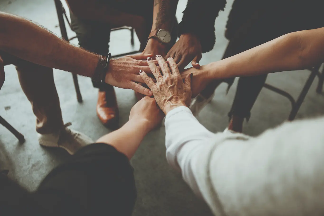 A group of people sitting in a circle place their hands together in the center, showing unity and teamwork. The image captures diverse hands overlapping on a gray surface.