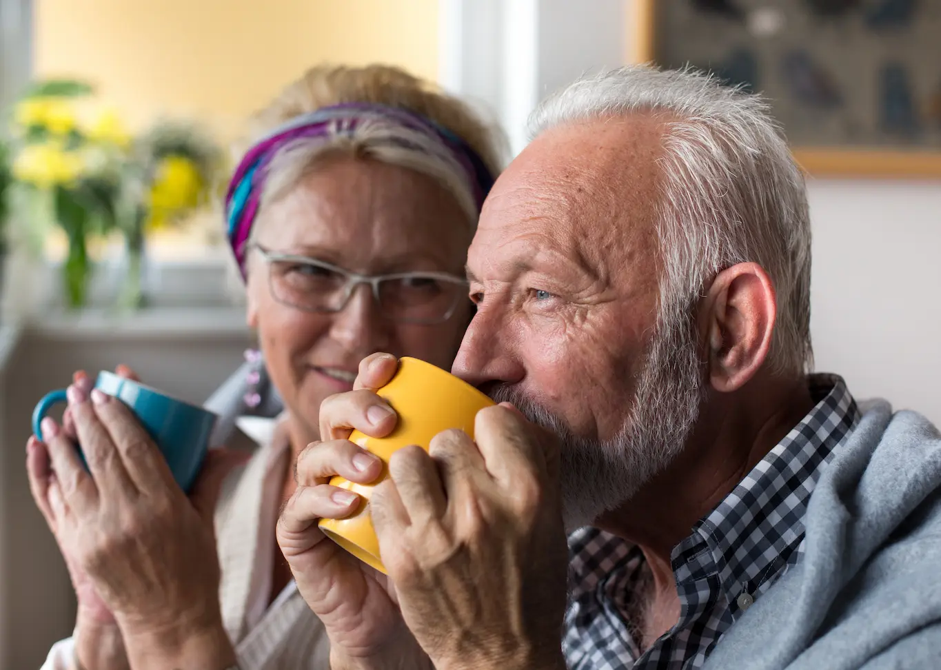 An older man with a gray beard and a checked shirt sips from a yellow mug, while an older woman with glasses and a headband smiles, holding a blue mug. They sit closely together in a cozy, sunlit room.