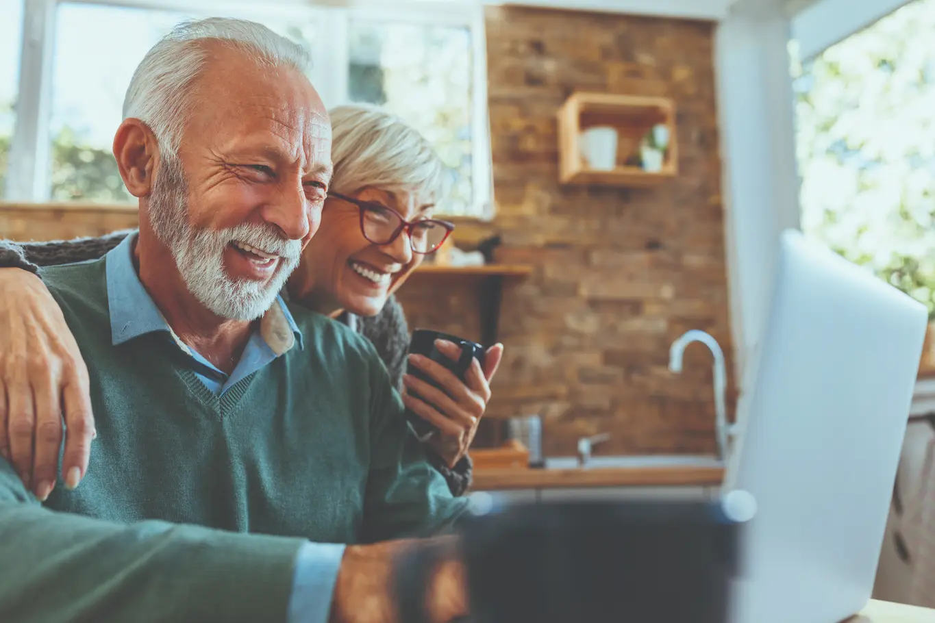 An older couple sits closely together at a kitchen table, smiling at a laptop screen. St.Louis Seniors can enjoy services like SeniorConcierge, making life easier as sunlight streams in and they savor coffee and connection.
