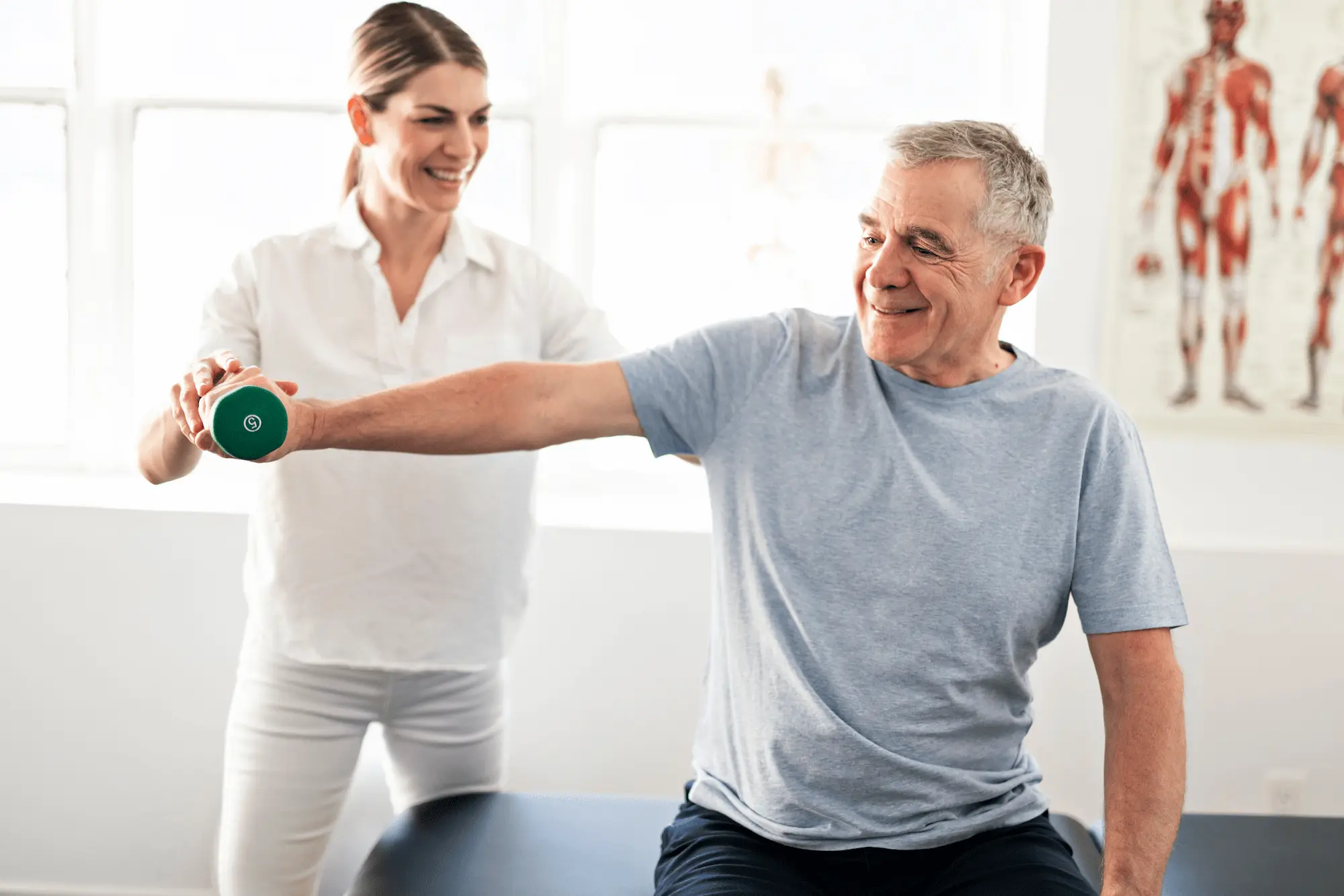A smiling older man lifts a small green dumbbell with his outstretched arm while a female physical therapist assists and encourages him in a bright clinic room.