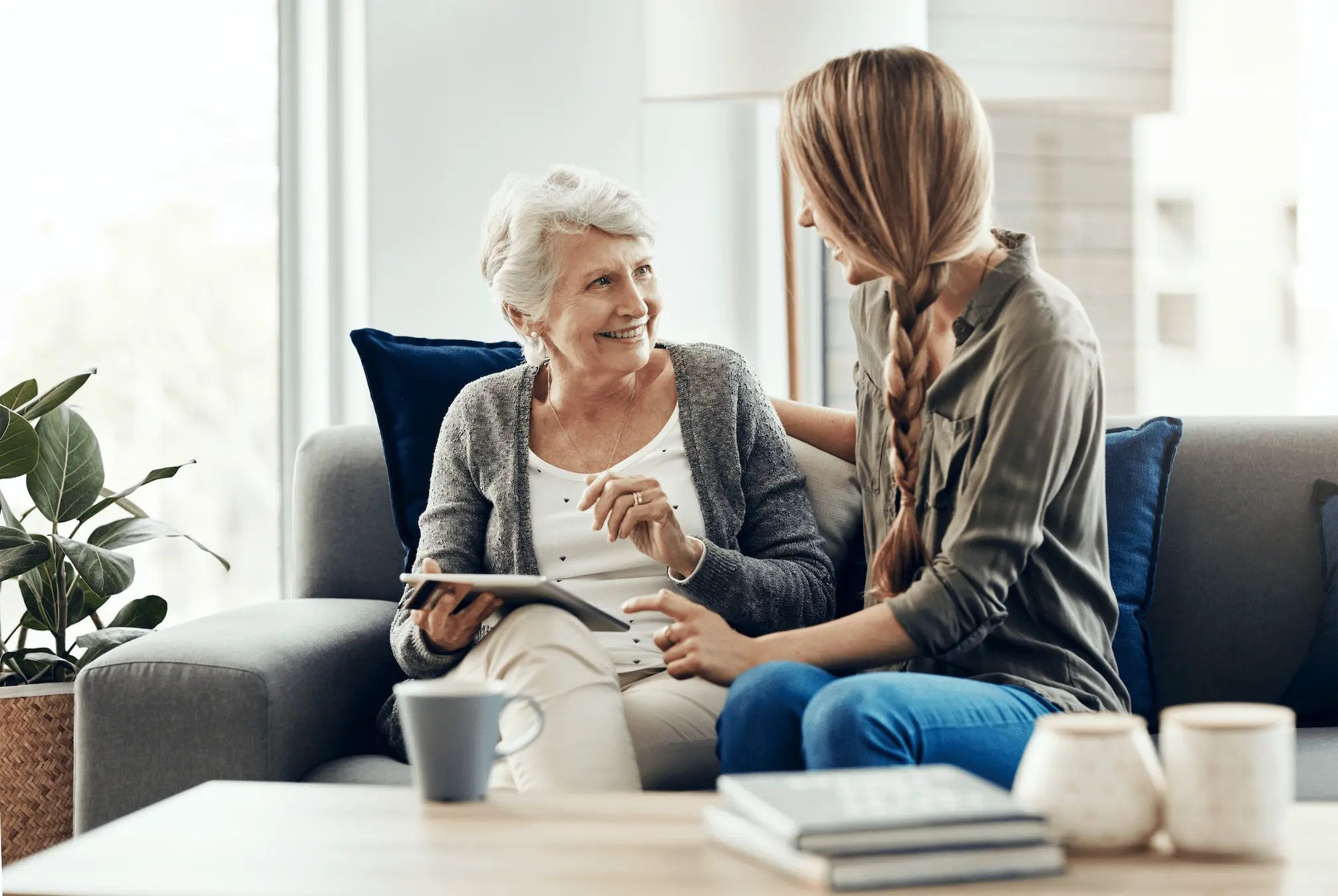 An older woman and a younger woman sit together on a couch, smiling and talking while looking at a tablet. They appear relaxed and happy in a cozy living room setting with coffee mugs and books on the table.