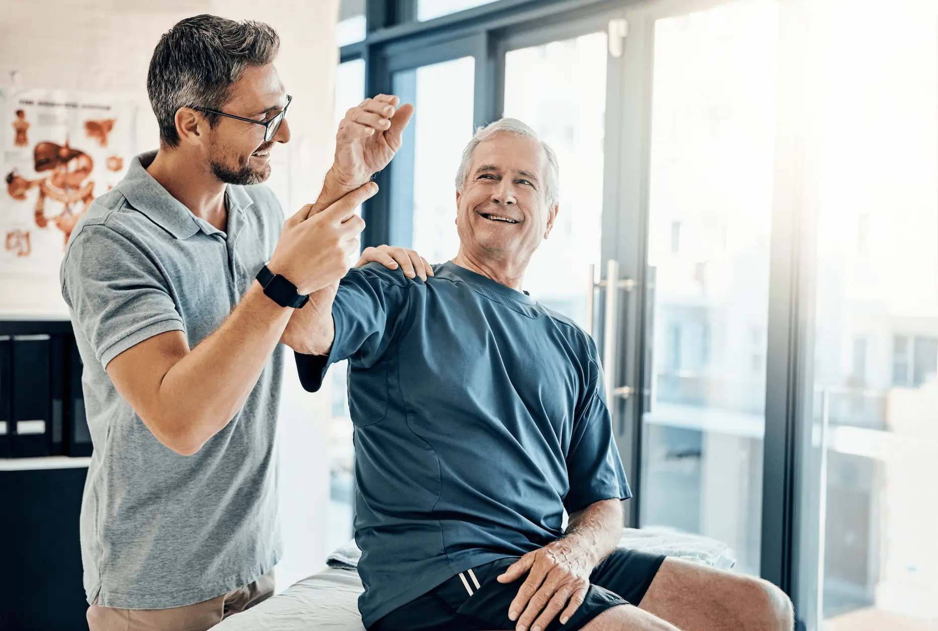 A smiling older man in athletic wear sits on an exam table while a healthcare professional helps him lift his arm, likely demonstrating a physical therapy exercise in a bright medical office.