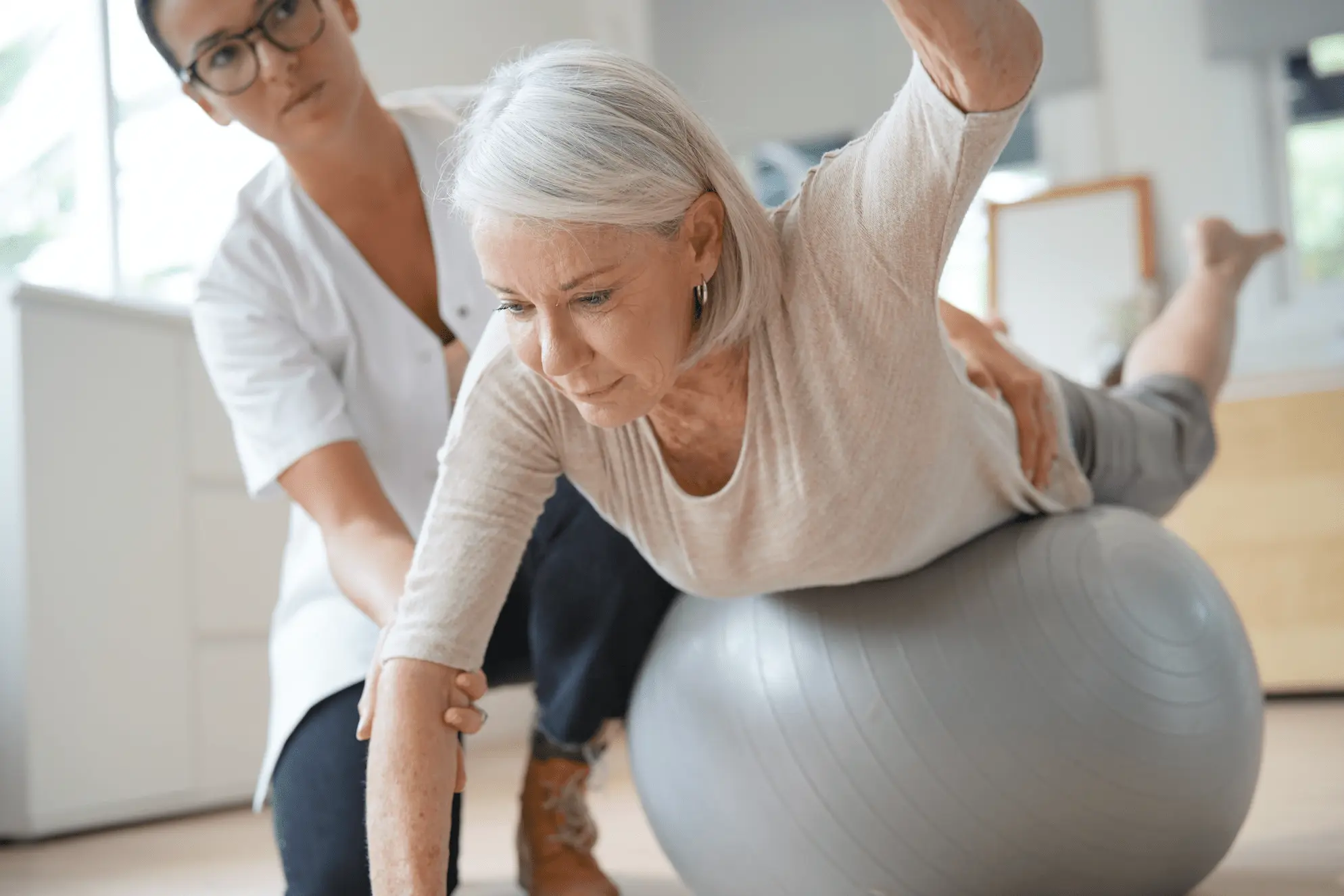 An elderly woman balances on a stability ball with her arms extended, while a female physical therapist supports and guides her in a bright rehabilitation room.