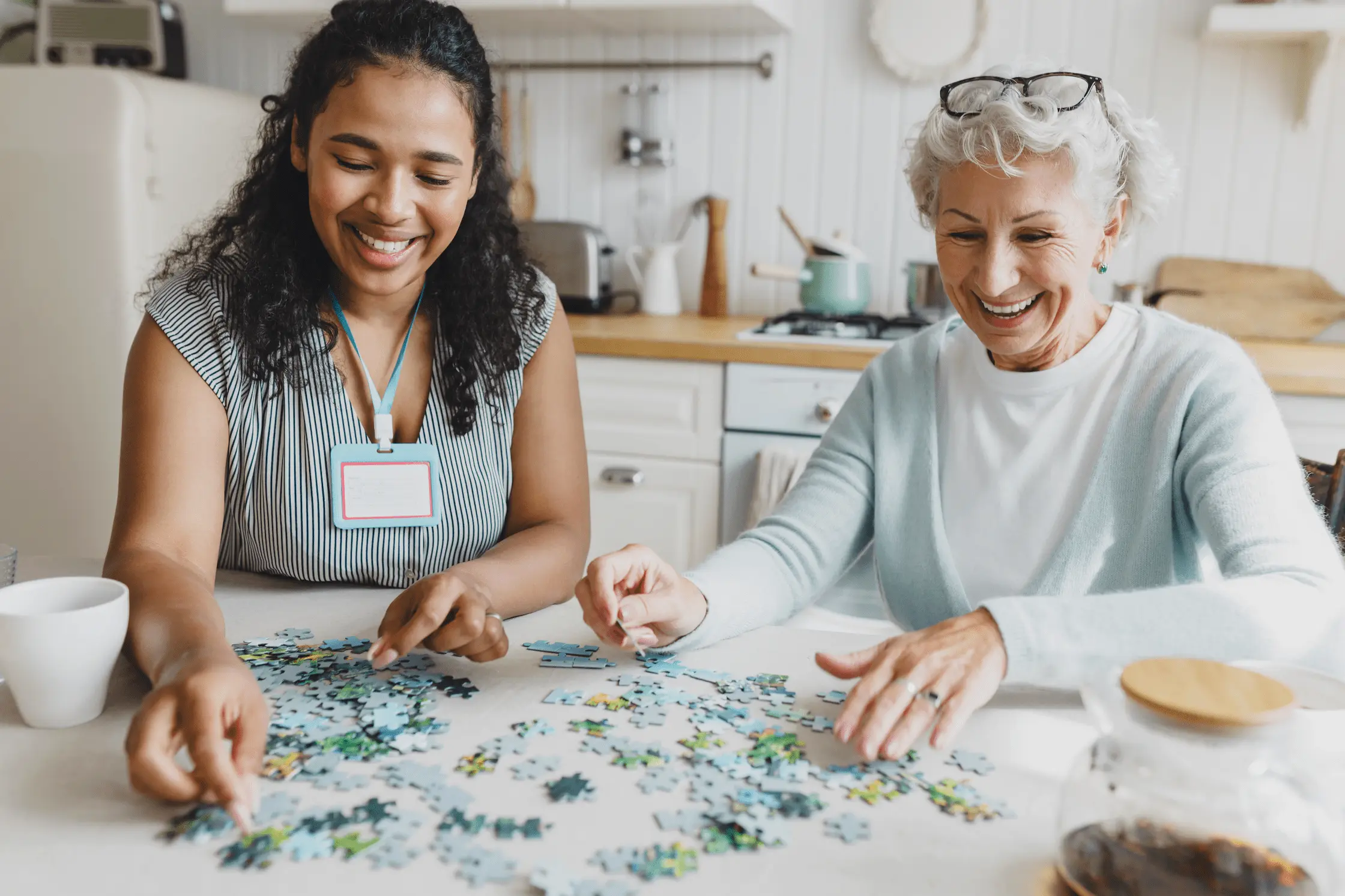 Two women, one younger and one older, smile and work together on a jigsaw puzzle at a kitchen table, appearing happy and engaged in the bright, cozy kitchen setting.