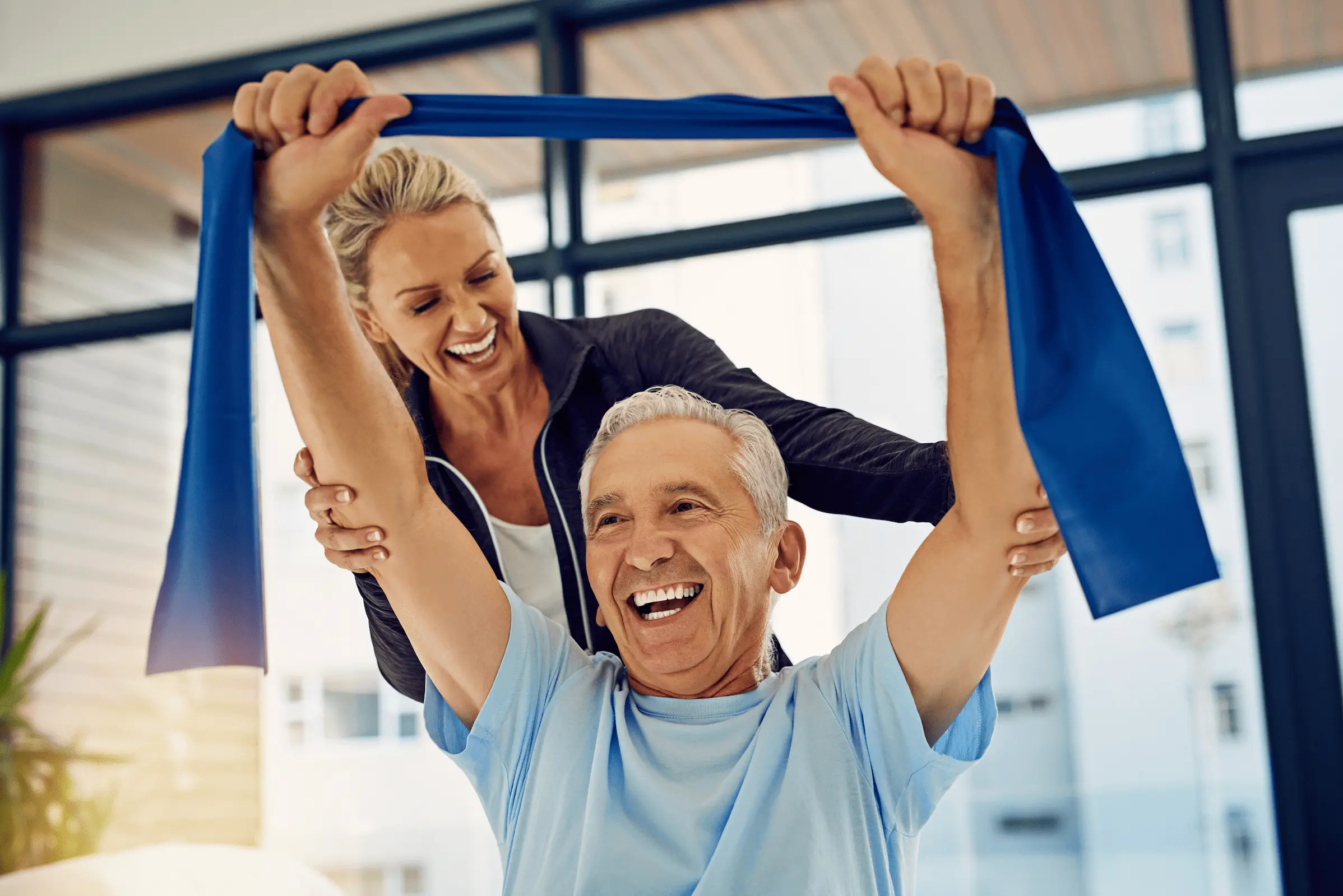 An older man smiles while lifting a blue resistance band overhead, assisted by a woman behind him in a bright room. Both appear happy and engaged in a fitness or physical therapy session.