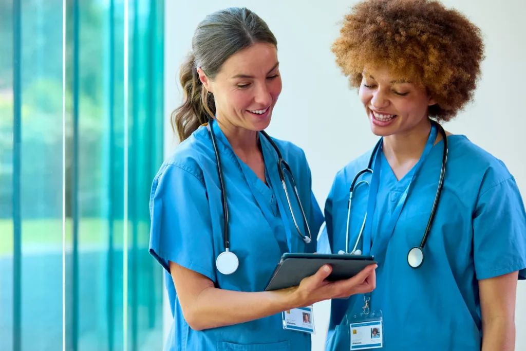 Two smiling healthcare professionals in blue scrubs and stethoscopes review information on a tablet together in a bright, modern medical facility.