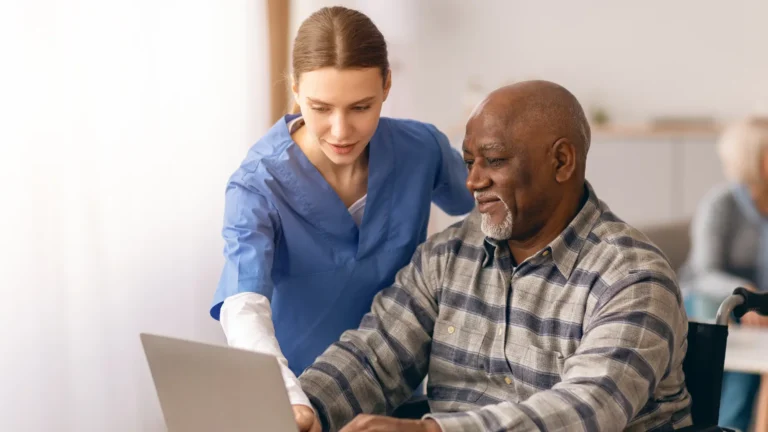 A nurse helps an elderly man in a wheelchair use a laptop. They both appear focused and engaged, sitting in a bright, indoor setting with another person blurred in the background.