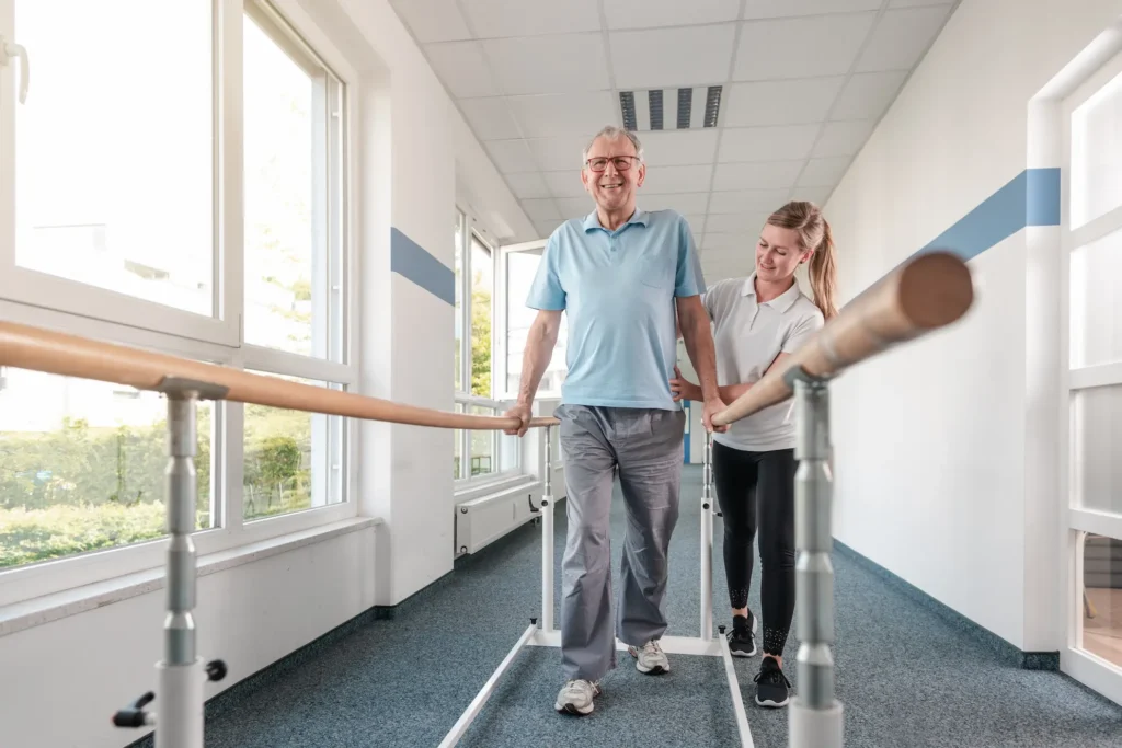 An older man walks between parallel bars in a bright rehab facility, assisted by a smiling female therapist. Sunlight shines through large windows.
